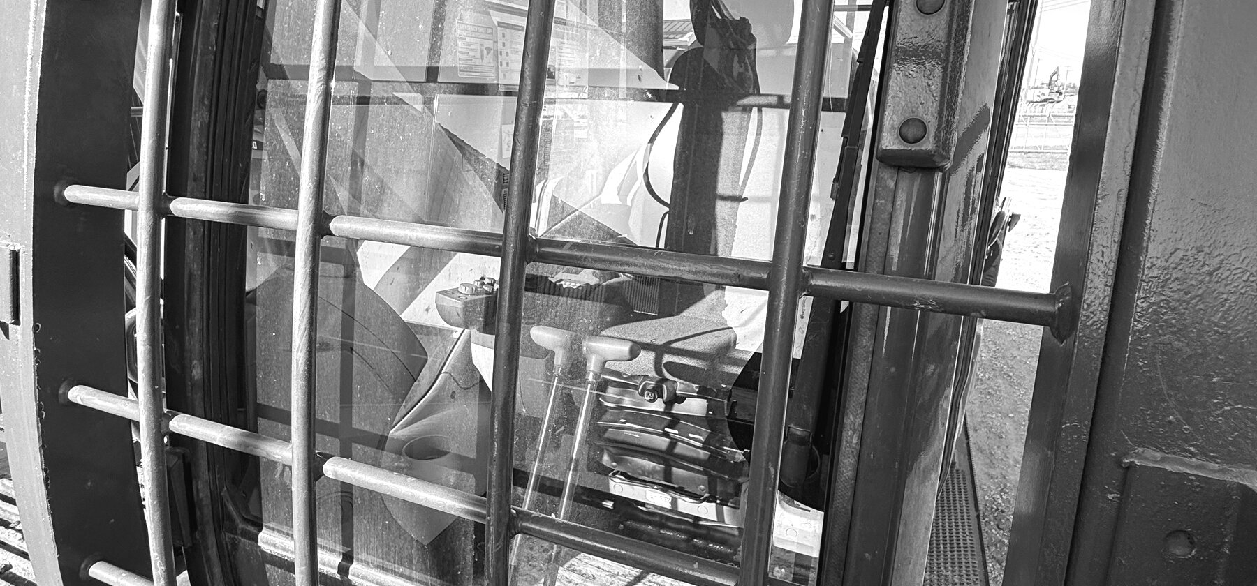 A steel window guard grille over an excavator cab door, the joystick and seat visible behind the glass