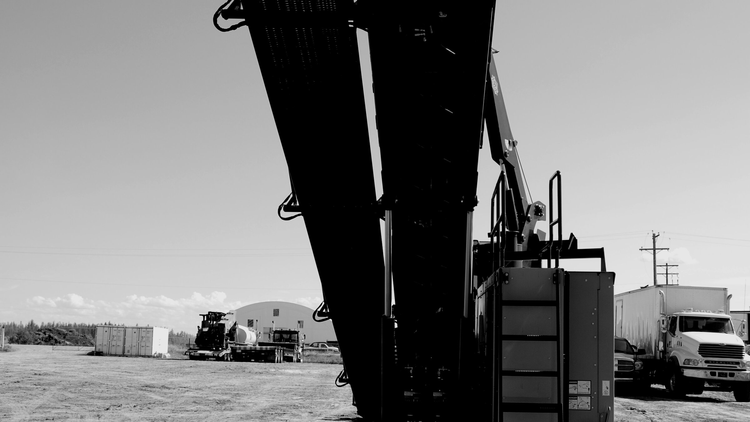 A drill rig's mast rising against the sky, low sun behind it, a yard of trucks and trailers below